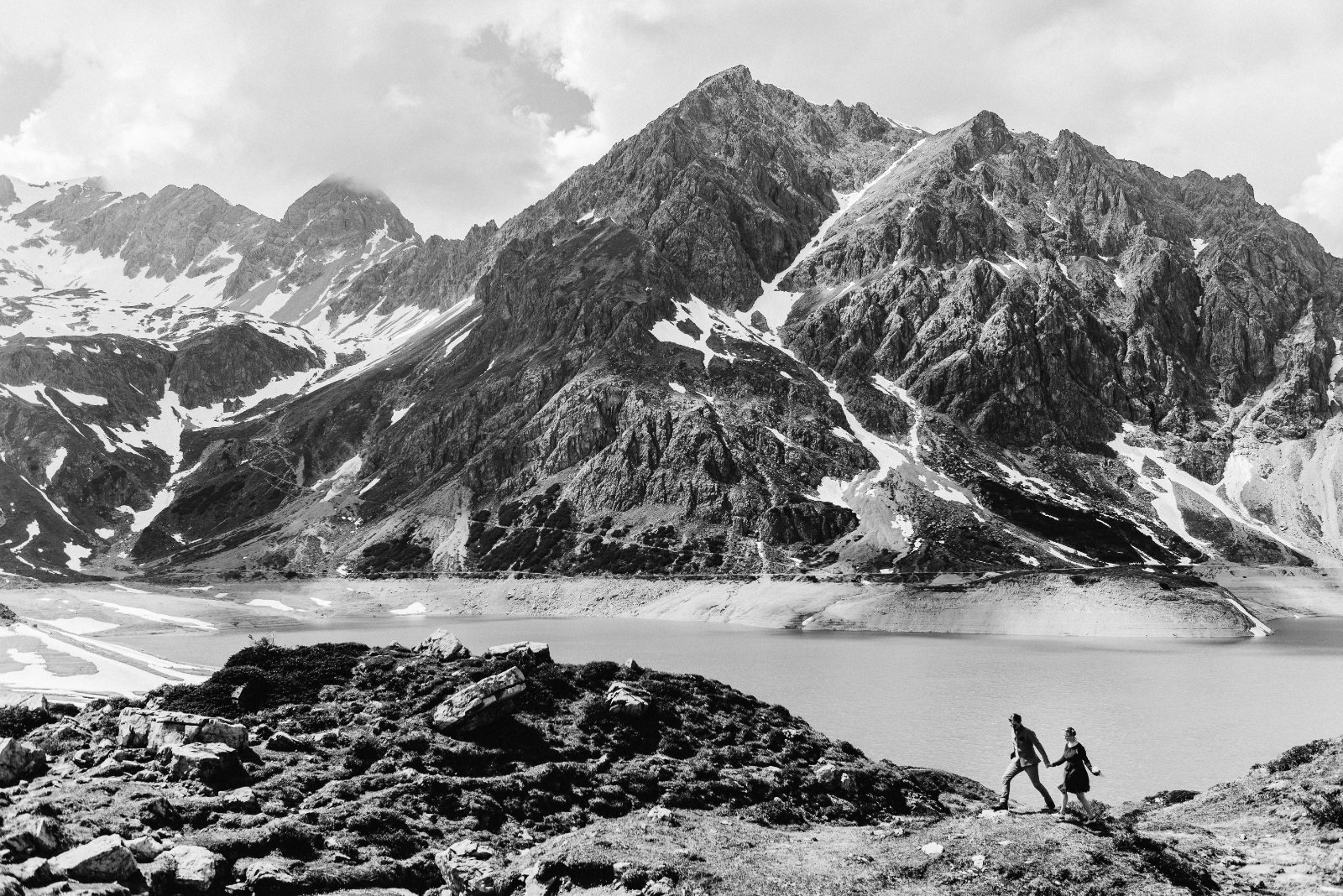 Brautpaar Hand in Hand auf einem Grat über dem Lünersee, dahinter der See und die verschneiten Gipfel — Schwarz-Weiß